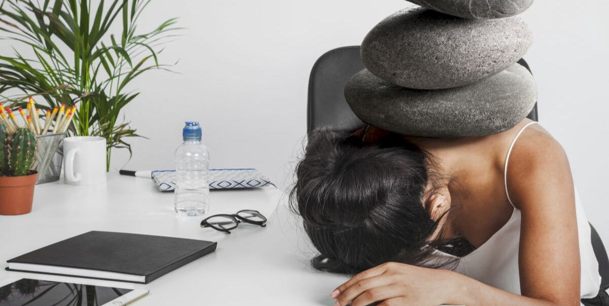 Persoană epuizată cu capul pe birou, având pietre mari așezate simbolic pe spate, sugerând stres și presiune la locul de muncă