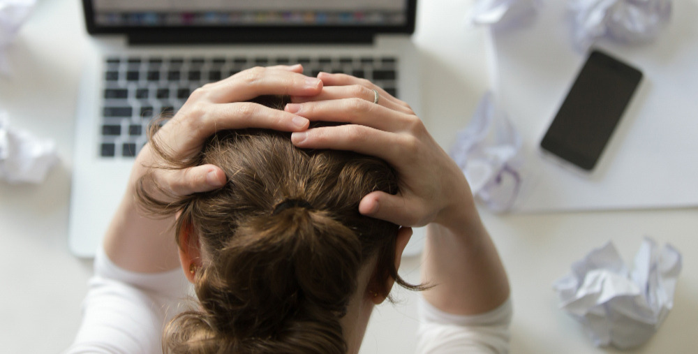 Portretul unei femei care se ține de cap la birou, lângă laptop.