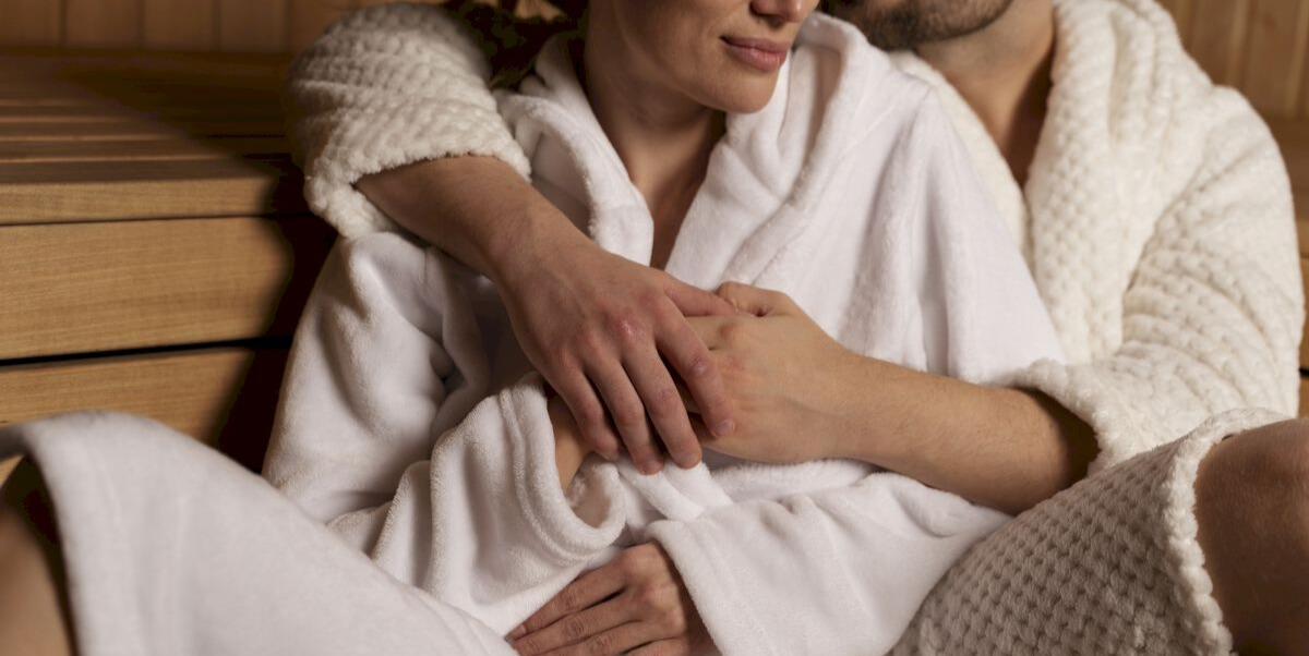 Cuplu îmbrăcat în halate albe, stând îmbrățișat într-o saună din lemn, într-un moment de relaxare.